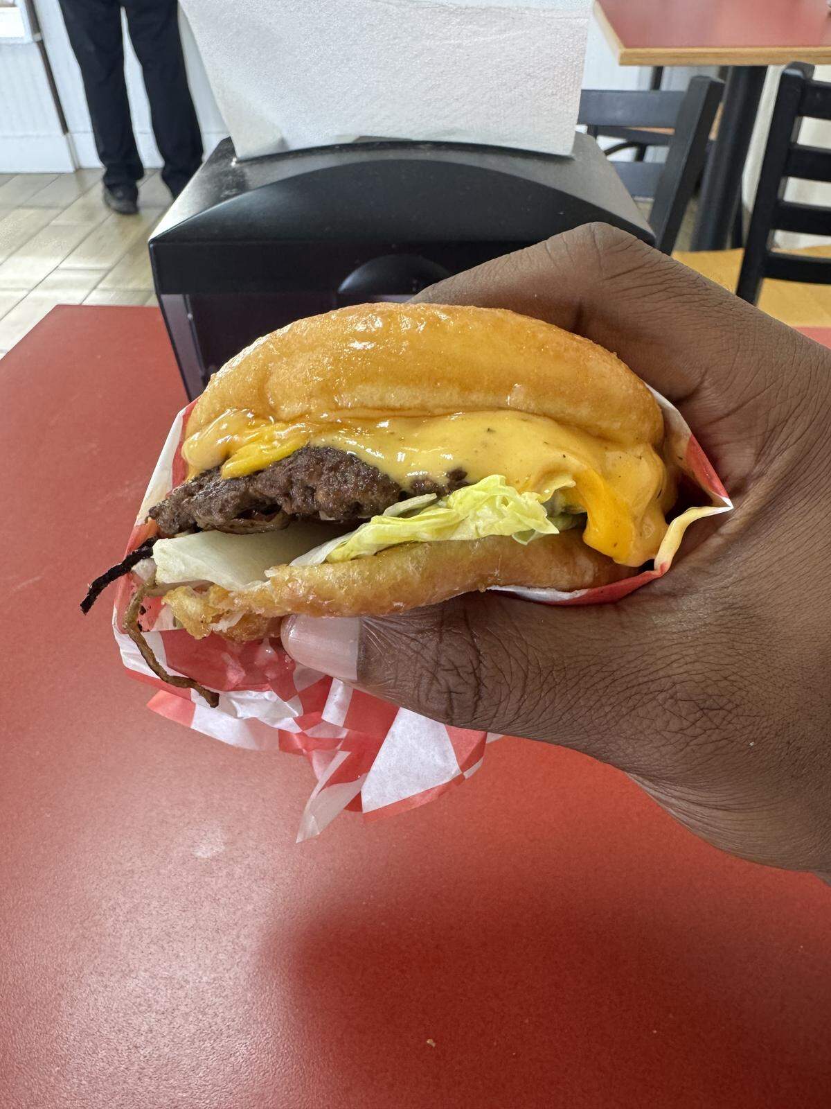 A close-up, first-person view of a doughnut burger being held in a hand. The burger consists of a savory meat patty, melted yellow cheese, and lettuce, sandwiched between two halves of a glazed doughnut instead of a traditional bun. It is wrapped in red and white checkered wax paper.
