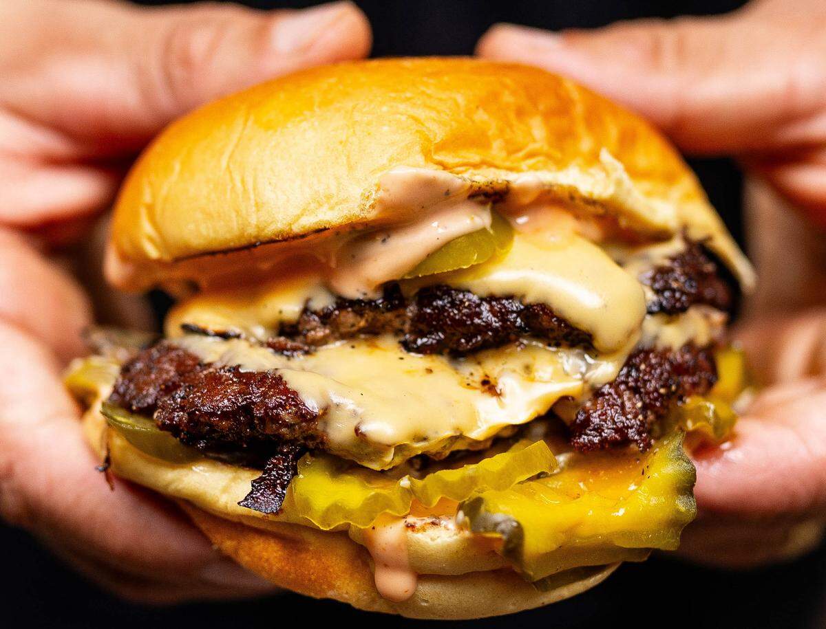 A detailed, close-up food photograph of a double smash burger being presented to the camera. The burger features two beef patties with a dark, lacy crust, layers of gooey melted cheese, sliced pickles, and a generous amount of a pinkish “secret sauce” on a toasted brioche bun.