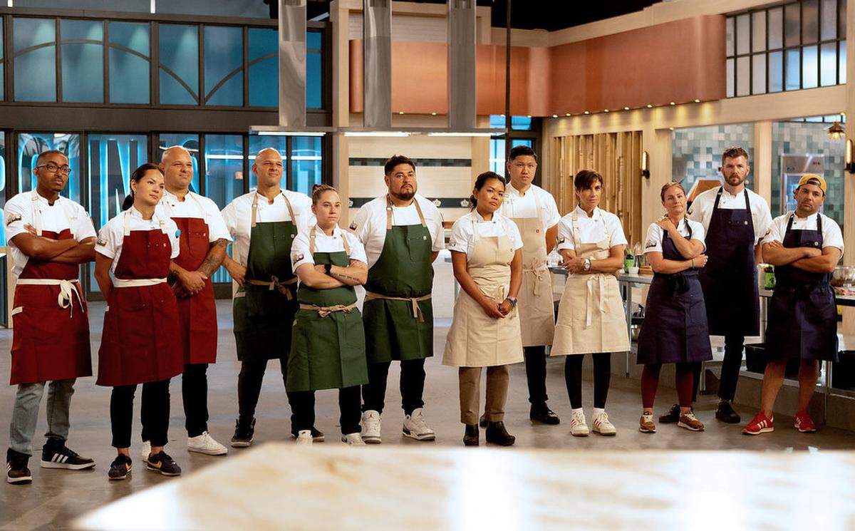 A wide shot of 11 chefs standing in a row on a professional kitchen set. They are all wearing white chef coats with various colored aprons, including burgundy, forest green, cream, and navy blue. Most have their arms crossed or hands behind their backs, looking toward the camera with serious, expectant expressions. The background features modern kitchen architecture with industrial lighting, wood paneling, and a large sign partially visible behind them.