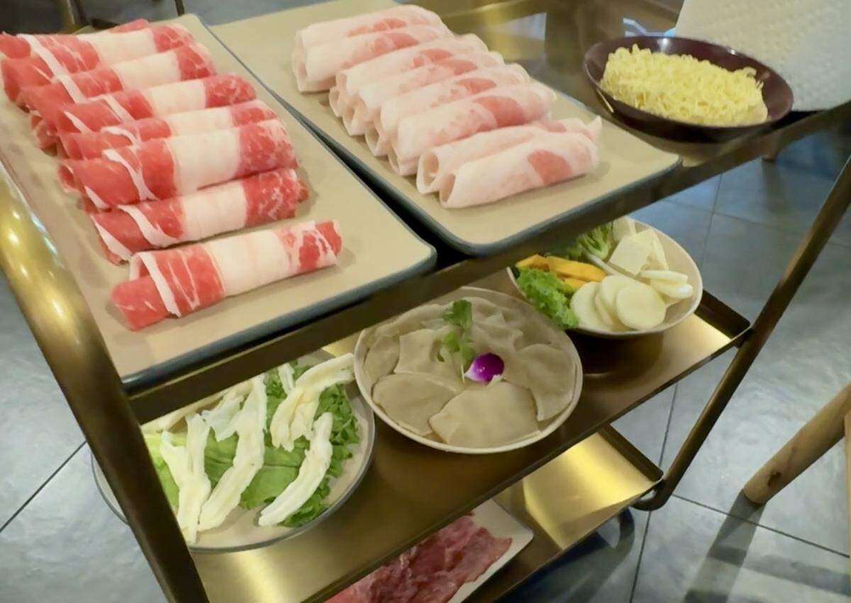 A three-tiered gold metal serving cart holding various hotpot ingredients. The top shelf features two large rectangular plates of thinly sliced, rolled raw meats (beef and pork) and a small bowl of yellow noodles. The lower shelves contain plates of leafy greens, sliced potatoes, tofu, and fresh dumplings.