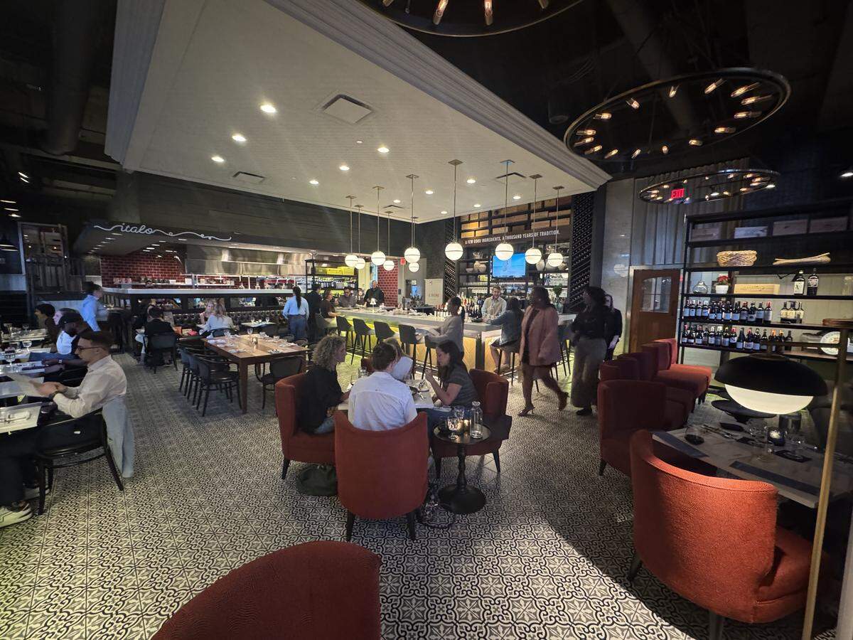 A wide, eye-level shot of a busy, upscale Italian restaurant dining room. The room features intricate black-and-white patterned tile flooring and warm orange armchairs arranged around small tables. In the background, a brightly lit bar area stands beneath a white dropped ceiling panel with recessed lighting and hanging white globe pendants. Large, circular wrought-iron chandeliers with exposed bulbs hang from the high industrial ceiling. Guests are seen dining, walking, and sitting at the bar.