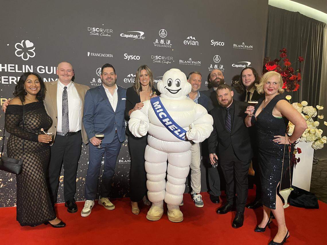 A group of people in formal and semi-formal wear, posing with the Michelin Man mascot on a red carpet. They are standing in front of a large black backdrop for the “MICHELIN GUIDE CEREMONY,” which features various sponsor logos. The group is smiling, and several people are holding drinks.