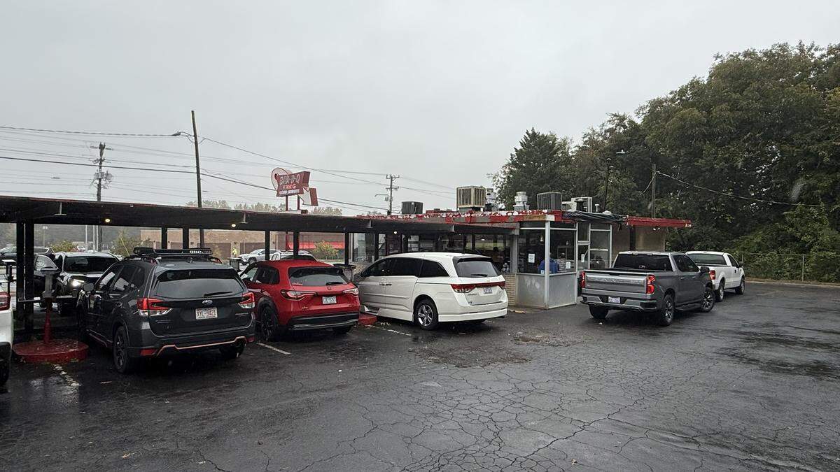 An exterior view of a drive-in style restaurant on an overcast day. Several cars, including SUVs and a pickup truck, are parked or waiting in a line that wraps around the building toward a covered service window. The parking lot is wet.