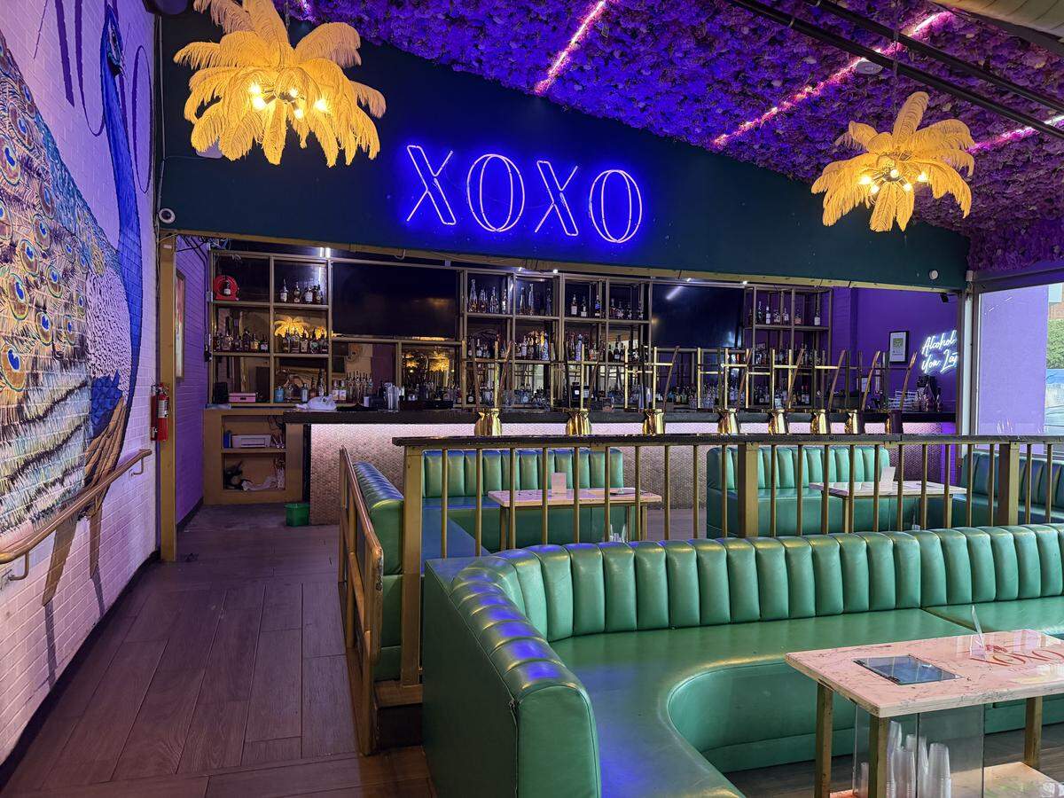 A wide shot of a brightly colored, stylish restaurant lounge featuring curved teal vinyl booths paired with light marble-top tables. In the background, a fully stocked bar sits under a glowing blue neon “XOXO” sign mounted on a dark green wall. The eclectic decor is highlighted by a dense ceiling arrangement of purple flowers, eye-catching gold palm-tree-shaped chandeliers, and a large peacock mural painted on a white brick wall to the left.