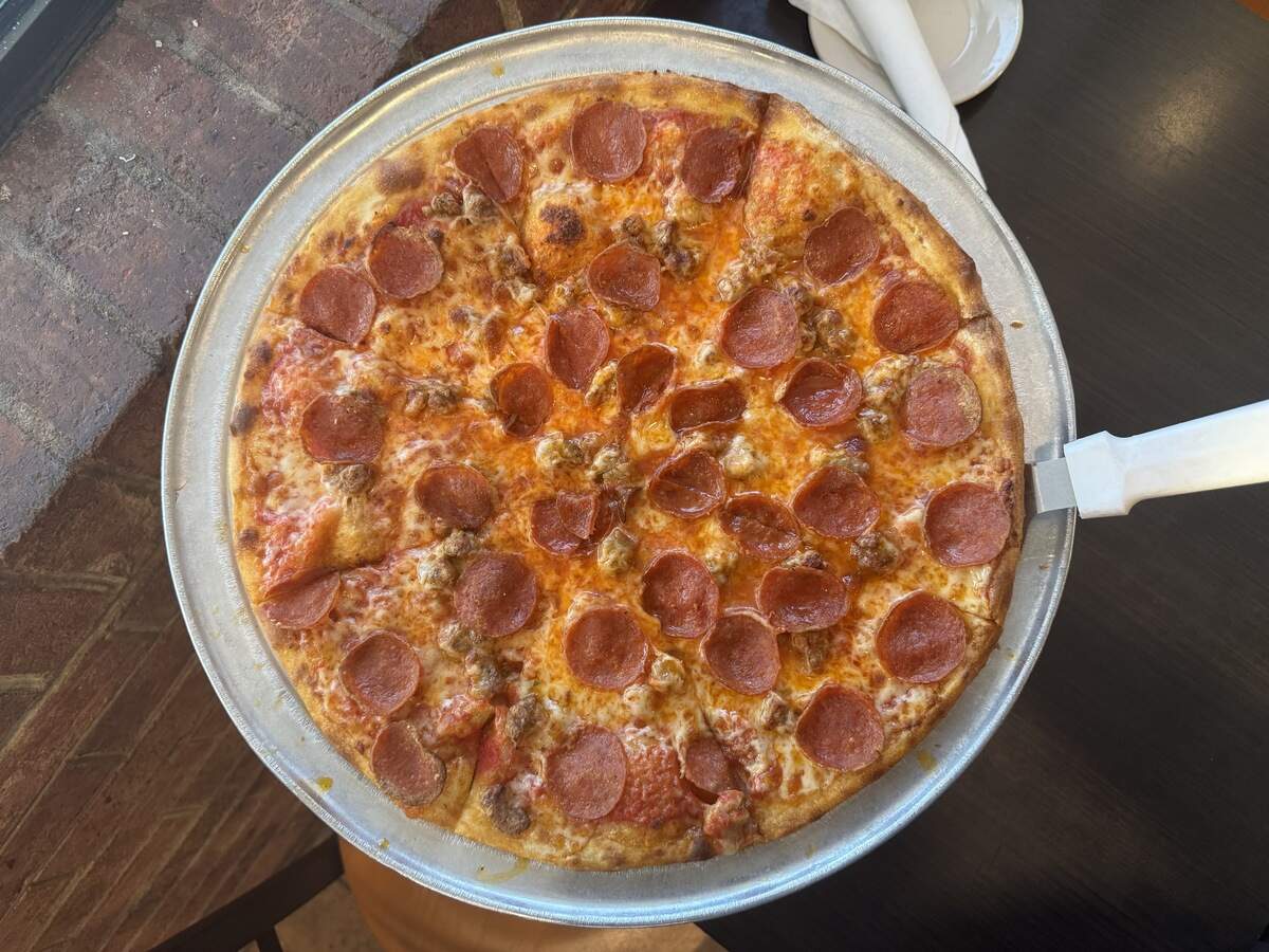 A top-down view of a whole, freshly baked pepperoni and sausage pizza served on a round, silver aluminum tray. The pizza features a golden-brown crust, melted orange-yellow cheese, scattered pieces of crumbled sausage, and numerous circular pepperoni slices. A white-handled pizza server is tucked slightly under the right side of the crust. The tray rests on a dark tabletop next to a textured brick wall ledge.