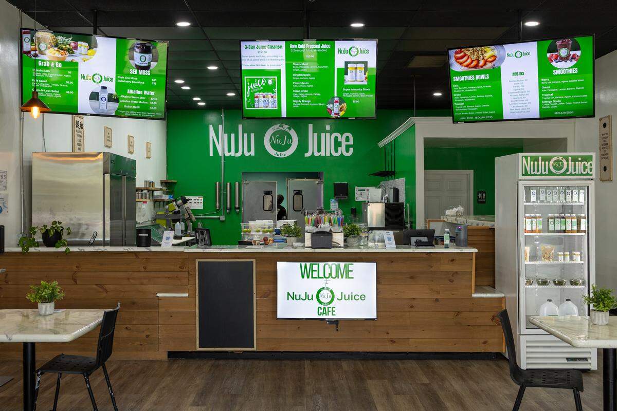 A wide, straight-on shot of the interior of the ‘NuJu Juice Cafe.’ The bright, modern space features a long, wood-paneled service counter in front of a green wall with the ‘NuJu Juice’ logo. Three large digital menu screens hang above, displaying items like juice cleanses and smoothies. A branded refrigerator full of bottled juices is on the right, and a few tables are in the foreground.