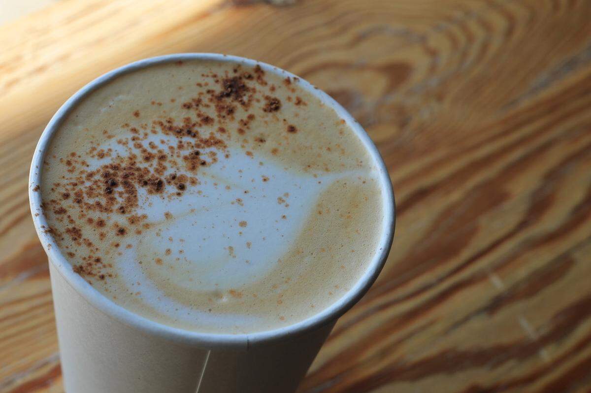 A top-down, close-up view of a paper coffee cup with a coffee drink topped with frothy milk and a light dusting of cinnamon or chocolate powder. The cup is sitting on a wooden tabletop with a noticeable woodgrain pattern.