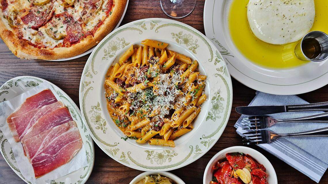 A vibrant, top-down photo of an Italian feast on a wooden table. In the center is a large plate of penne pasta in a creamy sauce, surrounded by a pepperoni pizza, a ball of fresh burrata in olive oil, sliced prosciutto and a bowl of roasted red peppers.