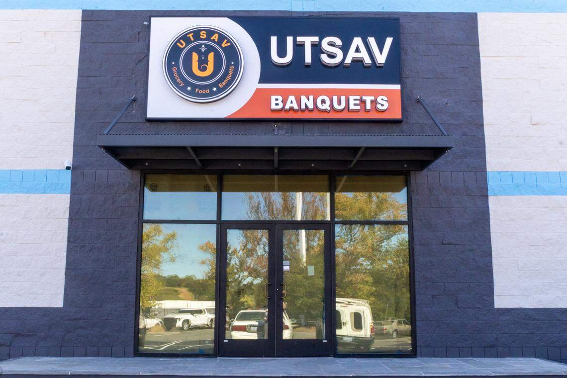 A close-up view of the Utsav Groceries, Food, and Banquets entrance is framed by dark gray brick-patterned walls. The double glass doors reflect the outdoor parking lot and a mix of trees and bushes in the background.