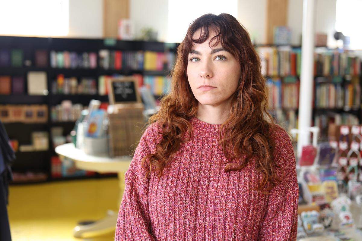 This image features a person with long, wavy reddish-brown hair standing in the center of a bookstore and wearing a textured, pinkish-maroon knit sweater. A neutral, direct expression is on the person’s face.