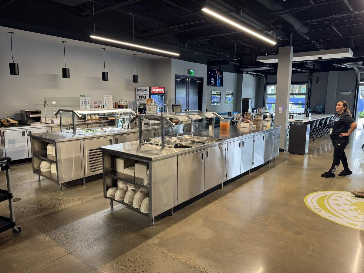 A wide shot of a modern, spacious cafeteria featuring long stainless steel buffet stations equipped with sneeze guards, plates, and bowls. The room has a polished concrete floor with a yellow circular logo in the foreground and sleek linear LED light fixtures overhead. In the background, long dining tables are arranged near large garage-style doors, and a staff member in black athletic wear stands to the right.