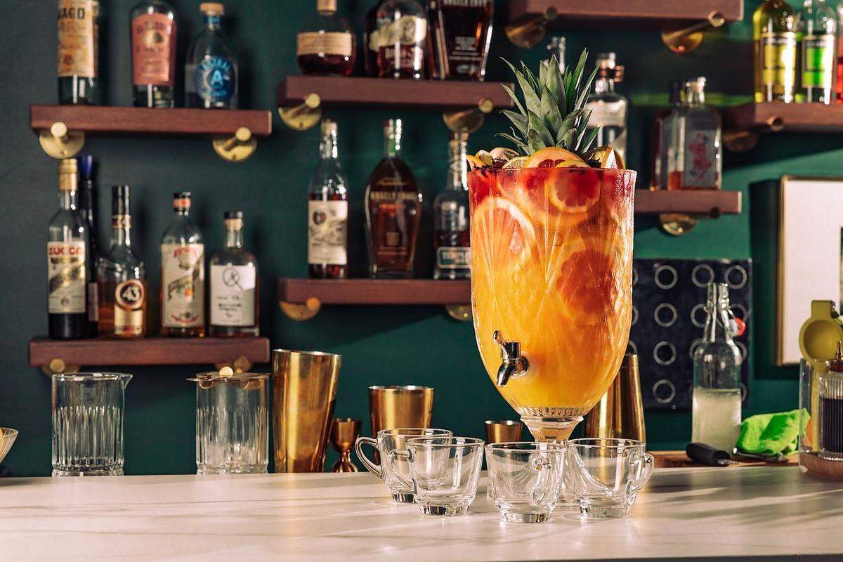 A stylish bar scene featuring a large, ornate glass beverage dispenser filled with a vibrant orange punch. The punch is garnished with slices of orange, grapefruit, and the top of a pineapple. Several small glass punch cups are arranged in front of the dispenser on a white countertop, with gold barware nearby. The background is a dark green wall with wooden shelves displaying a collection of liquor bottles.