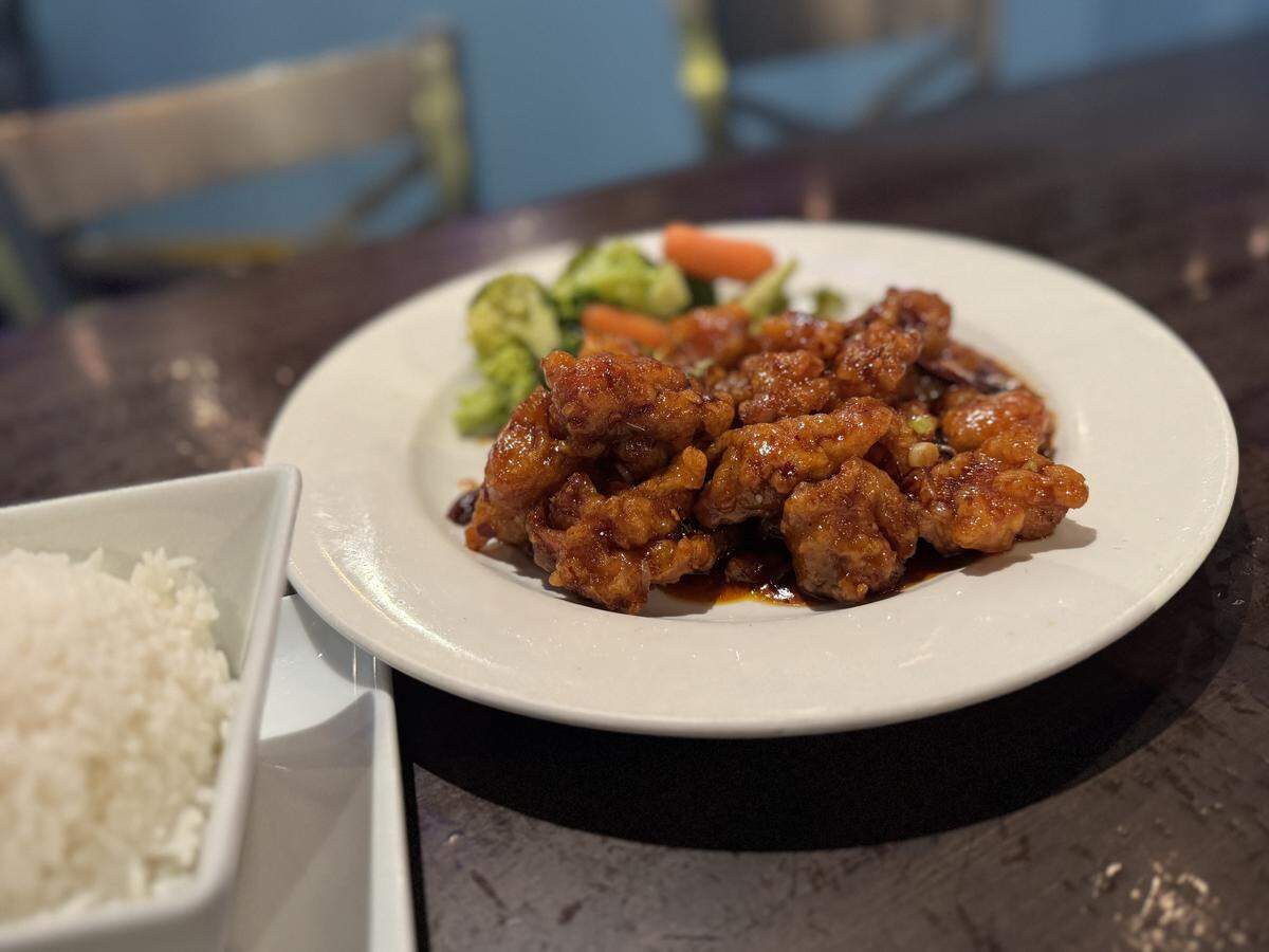 A dish of General Tso’s chicken from Baoding restaurant sits on a dark table. The crispy chicken is coated in a rich, dark sauce and is served on a white plate with steamed vegetables. A bowl of fluffy white rice is in the foreground, and the restaurant interior is blurred in the background.