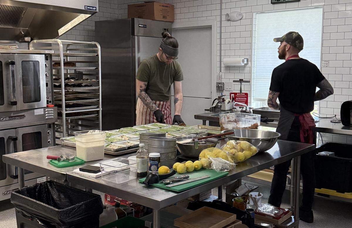 Two chefs working in a commercial kitchen with white subway tile walls. They are preparing several rectangular containers of food on a large stainless steel prep table.