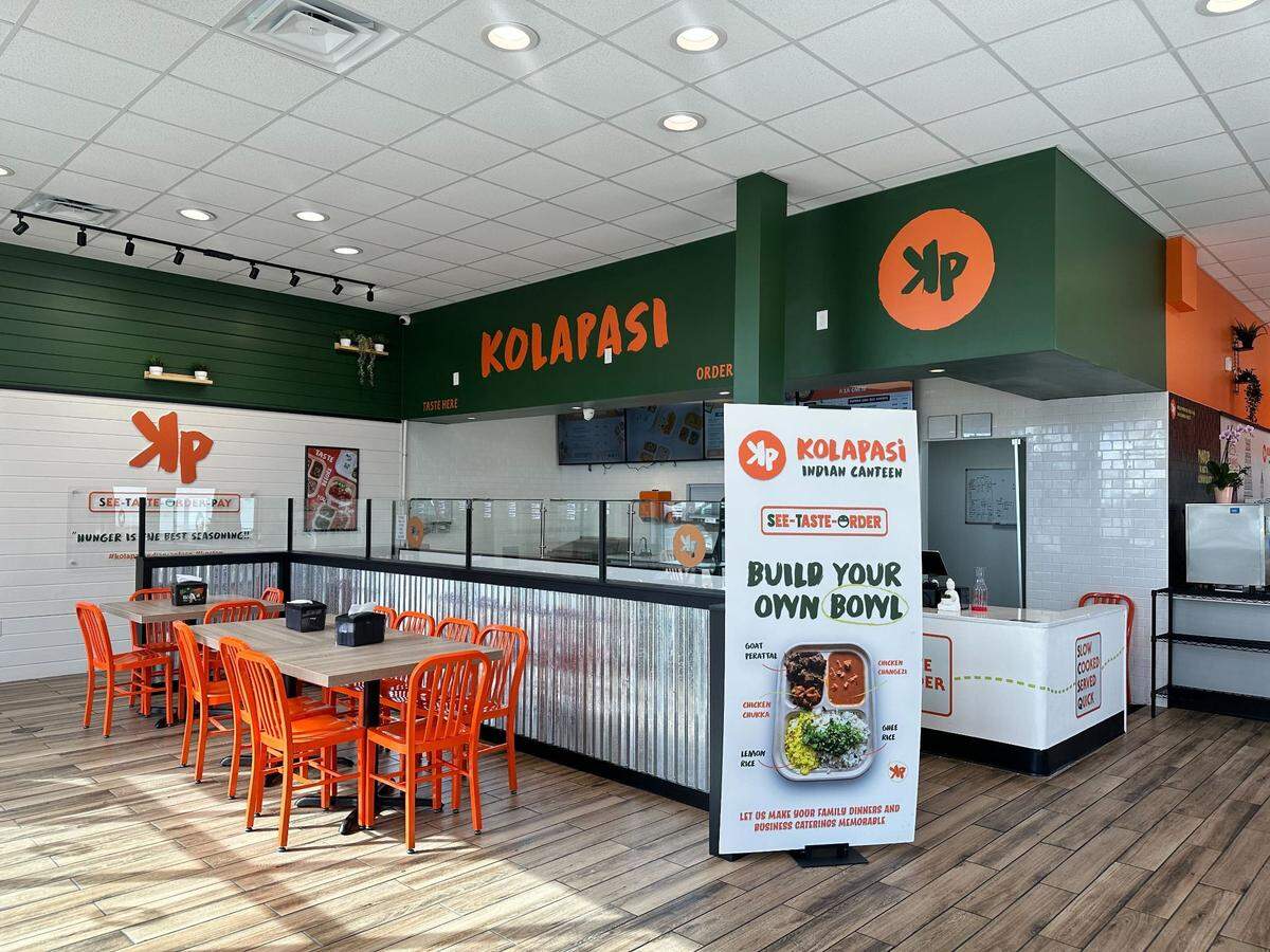 The interior of Kolapasi Indian Canteen features a modern design with green and orange walls, bright orange chairs, and wood-style flooring. A large white banner in the foreground promotes a “Build Your Own Bowl” option with a photo of a sample meal.