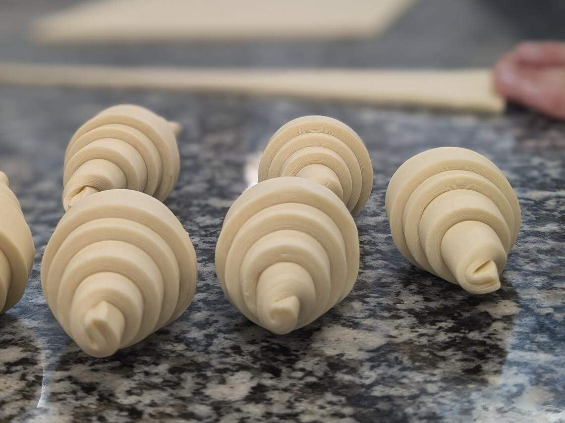 Several pieces of raw croissant dough are tightly rolled into their classic crescent shapes and arranged on a marble counter. In the blurred background, a baker’s hands are visible, working on stretching out a large sheet of dough.
