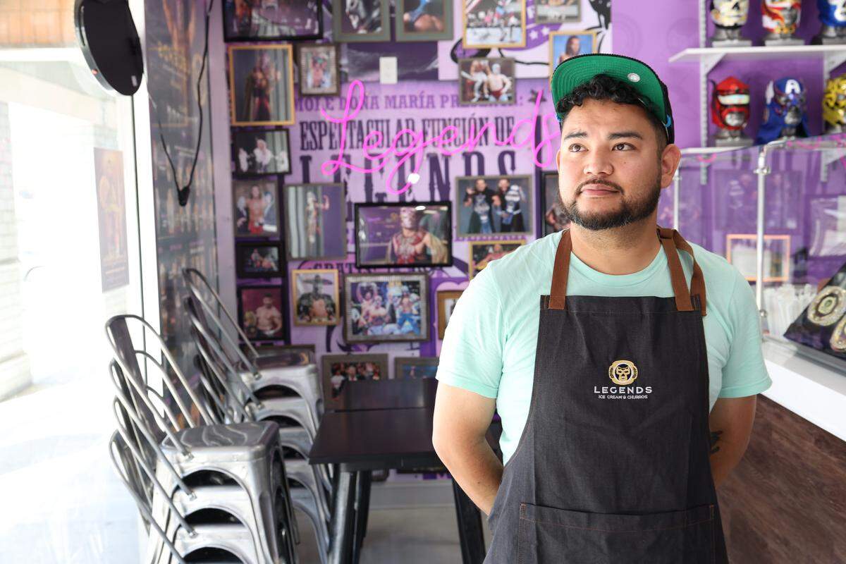 An eye-level, medium shot of a business owner wearing a mint-colored t-shirt, a black apron, and a backward green baseball cap, standing inside a restaurant. The restaurateur is looking off to the side, in front of a purple wall covered with framed photos of professional wrestlers, a pink neon sign, and shelves displaying colorful luchador masks. To the left, metal chairs are stacked next to a window.
