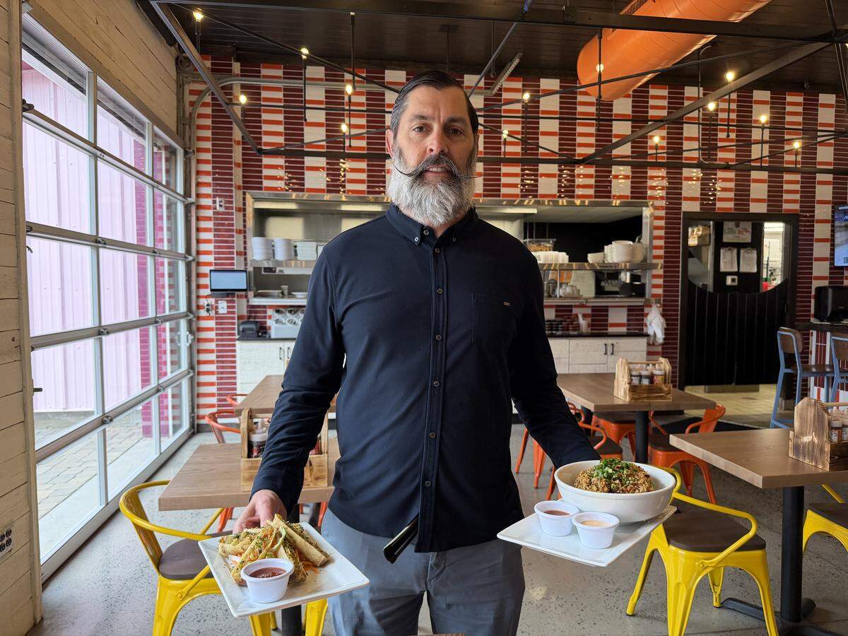 A restaurateur with a well-groomed, long grey beard and a styled mustache stands in a modern restaurant, holding two plates of food and wearing a dark navy button-down shirt and grey trousers. With the right hand, the restaurant owner holds a plate of crispy eggrolls with a side of red dip; with the left, a large bowl of fried rice garnished with green onions and two small dipping cups. The restaurant background features red and white striped wall tiles, industrial lighting, and bright yellow and orange chairs.