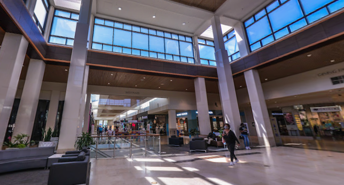 A wide-angle, low-level interior view of a modern, multi-story shopping mall atrium, notable for its high ceilings, natural light, and contemporary design. The space features large pillars, multiple levels of storefronts visible down the main corridor, and expansive windows near the ceiling that let in bright blue sky and sunlight.