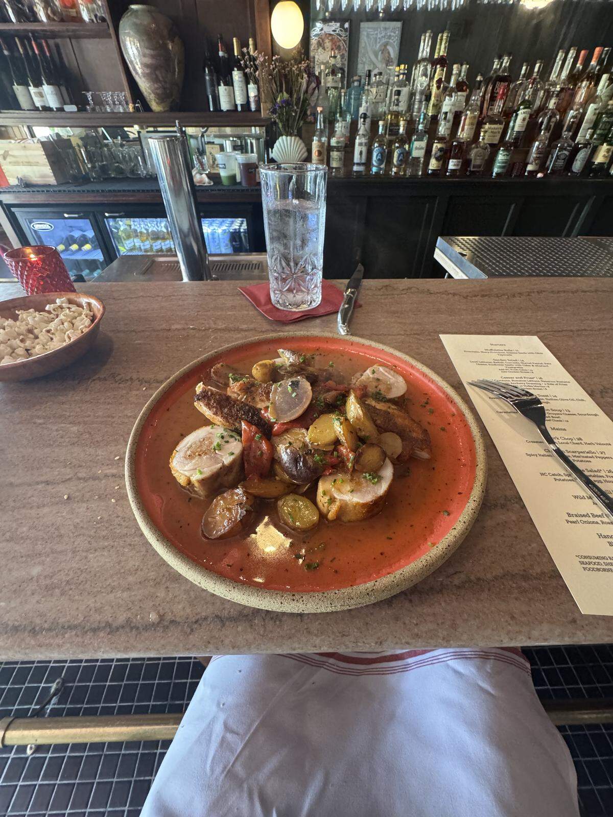 A first-person perspective of a meal served at a bar. In the center is a rustic orange plate containing sliced roasted chicken, potatoes, and peppers in a savory sauce. A glass of water, a small bowl of popcorn, and a menu are on the stone bartop next to the plate.