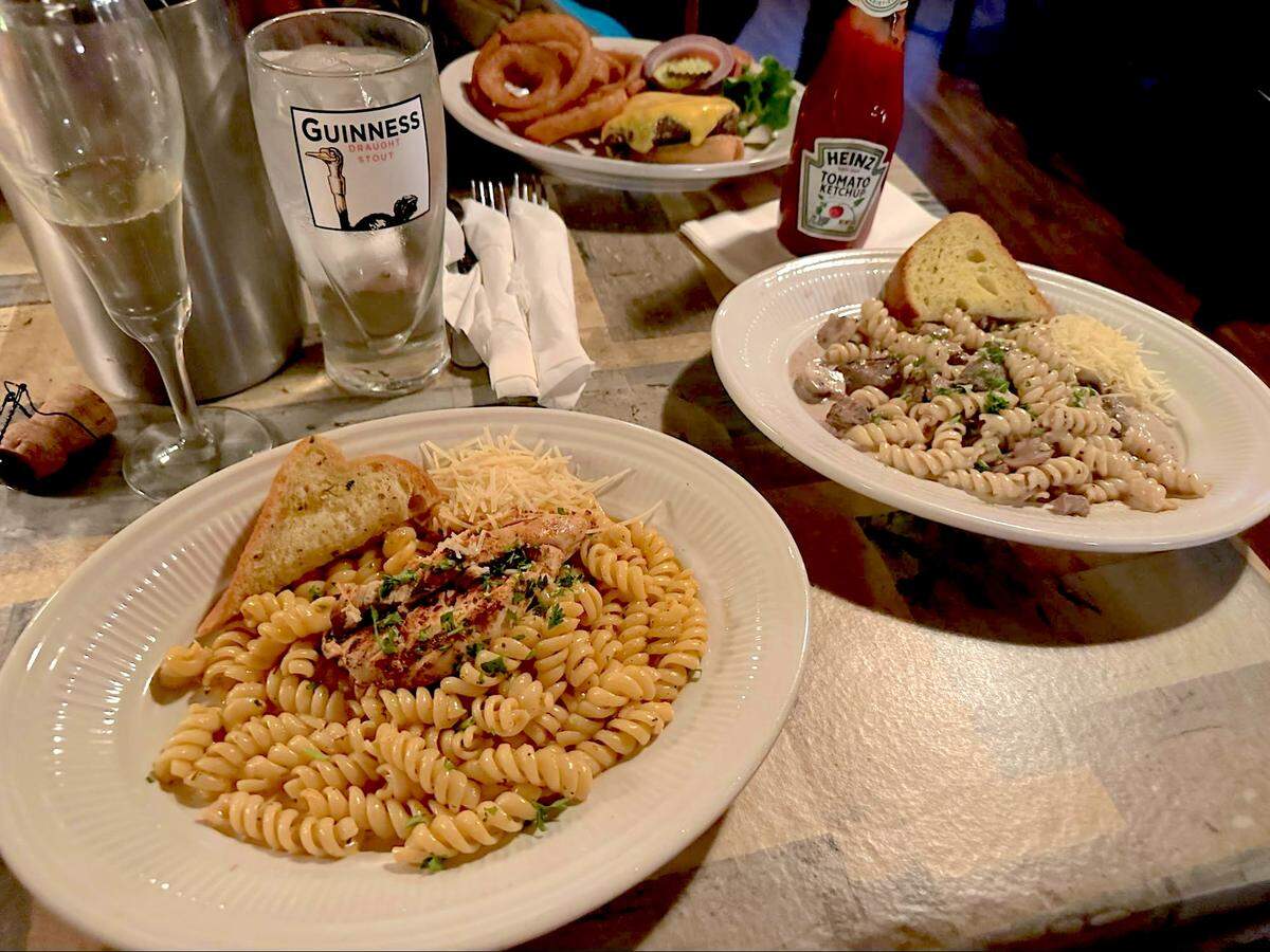 A slightly elevated view captures a casual dinner spread on a rustic wooden table. In the foreground, two white oval plates are prominently displayed. The plate on the left holds a serving of fusilli pasta topped with a piece of chicken, garnished with herbs and grated cheese, accompanied by a piece of toasted bread. The plate on the right features a similar pasta dish with mushrooms and what appears to be a sausage, also with a slice of bread and grated cheese. In the mid-ground, a tall Guinness draught stout glass is filled with water, next to a champagne flute with a bubbling drink. Further back, another plate is visible with a burger topped with cheese, lettuce, and tomato, served with a side of onion rings. A red bottle of Heinz Tomato Ketchup stands upright near the burger plate.
