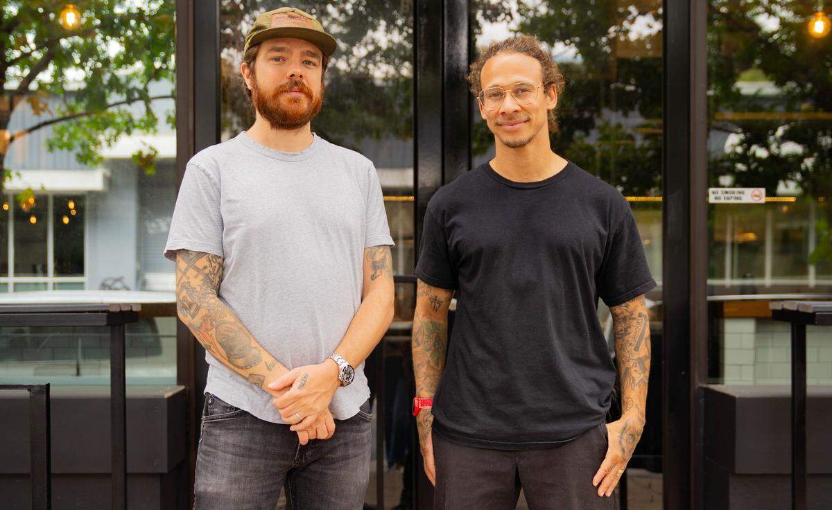 A side-by-side portrait of two people. On the left, Phillip Frankland Lee, with a red beard and wearing a cap, looks at the camera. On the right, Neen Williams, with curly hair and glasses, smiles slightly. They both have extensive arm tattoos and are standing in front of the glass entrance to a building.