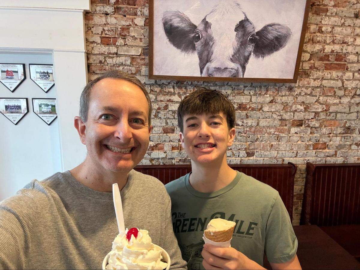 A selfie of a person and a younger person smiling inside the shop. The one on the left holds a cup of ice cream topped with a mountain of whipped cream and a cherry, while the younger one on the right holds a waffle cone. A large painting of a cow’s face hangs on the brick wall behind them.