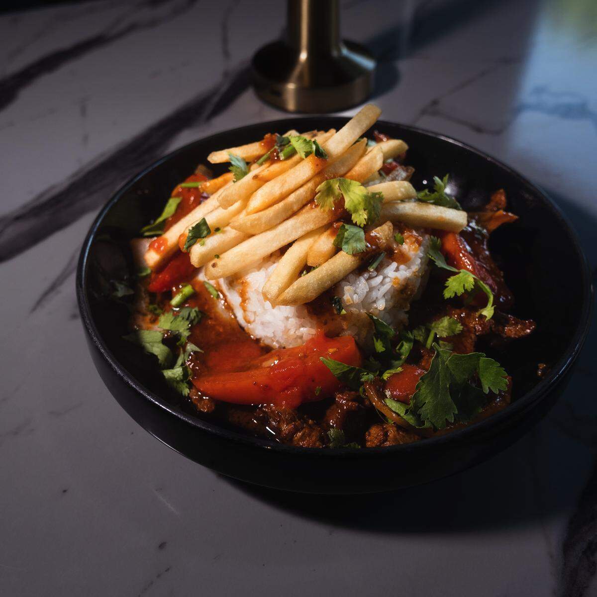 A dramatic, close-up shot of a Lomo Saltado dish in a black bowl. A mound of white rice and French fries is at the center, surrounded by a rich brown and red stew of beef and vegetables. The dish is garnished with fresh green cilantro.