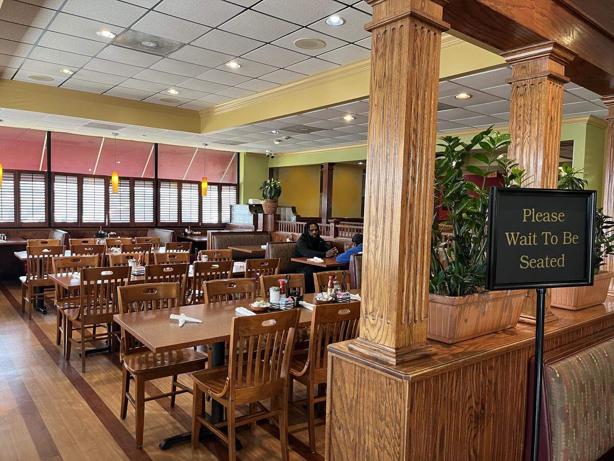 The interior of a spacious, well-lit restaurant dining room featuring rows of wooden tables and chairs, a banquette along the walls, and tall wooden columns. A small “Please Wait to Be Seated” sign rests near a column on the right.