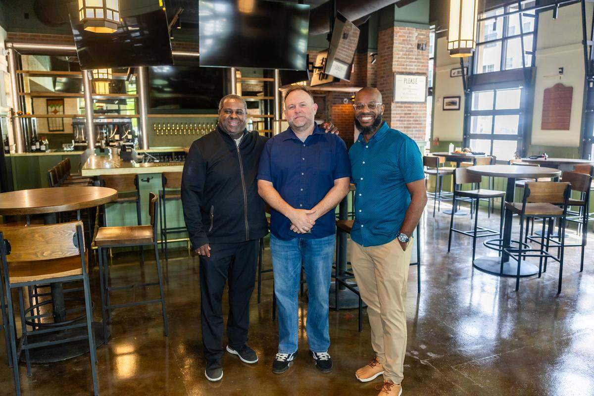 Three restaurateurs stand together smiling in the center of the pub’s bar area. The one on the left wears a black zip-up jacket, the one in the center wears a short-sleeved navy button-down and jeans, and the one on the right wears a teal patterned shirt and tan chinos. The background shows the modern bar with gold-toned countertops, multiple wall-mounted TVs, and industrial-style lighting.