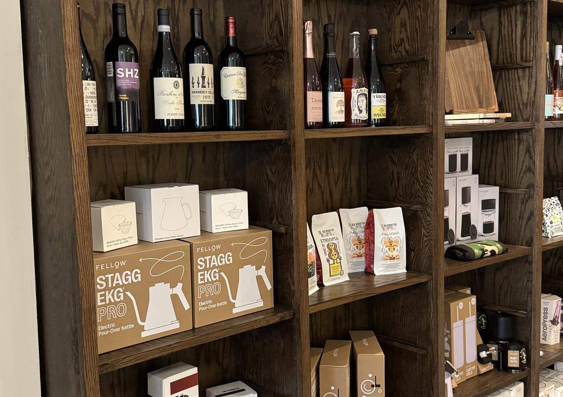 A close-up view of dark wooden shelving displaying retail goods, suggesting a cafe or gourmet retail section. The top shelves are stocked with bottles of red and sparkling wine. The middle shelves feature coffee-related products, including packaged coffee beans from Ethiopia and Nicaragua, and boxes for Fellow Stagg EKG electric pour-over kettles and other brewing equipment.