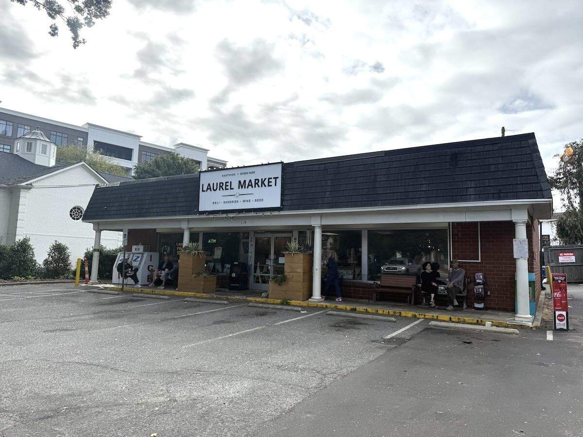 The exterior of Laurel Market, a single-story neighborhood deli with a red brick facade and a dark shingled roof, is seen from across its parking lot on an overcast day. A large white sign with the market’s name is prominent above the entrance. A few patrons are sitting on benches and planters along the front of the store under a covered walkway.