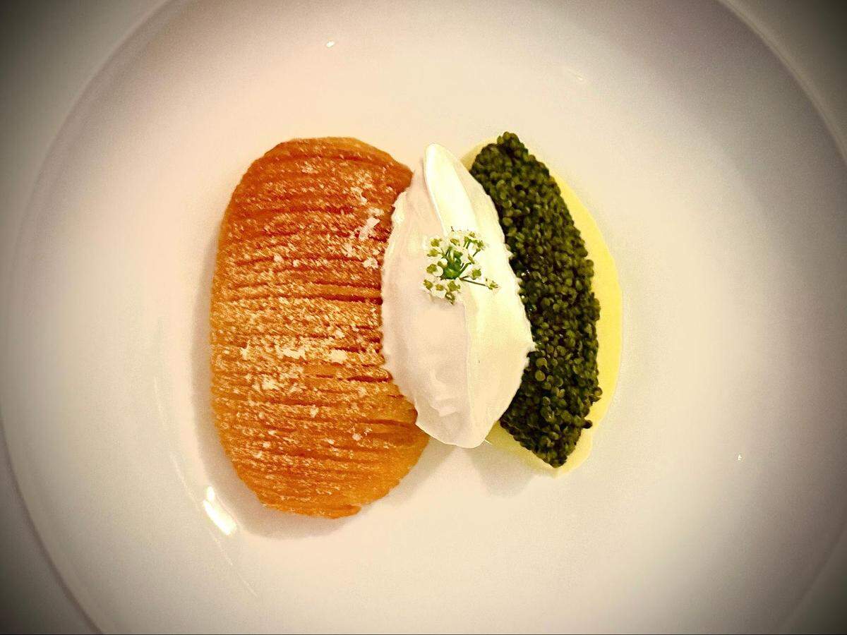 An overhead, close-up shot of a white plate with three components elegantly arranged in the center. On the left is a golden-brown, oval-shaped hasselback-style potato. In the middle is a smooth, white quenelle, possibly sour cream, garnished with tiny white flowers. On the right is a generous serving of dark caviar, resting on a base of what appears to be a light-colored purée.