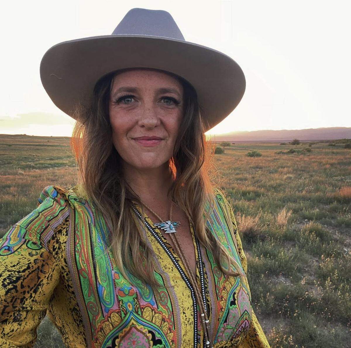 A person with long wavy hair and freckles smiles at the camera, wearing a light-colored fedora-style hat, and a vibrant yellow and green patterned dress. The individual is standing outdoors in a dry grassy field or prairie at sunset, with the golden light creating a warm rim-light effect.