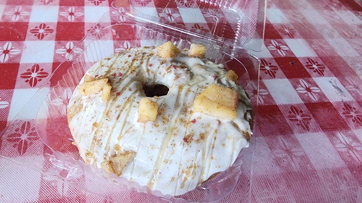 An overhead shot of an Apple Cider Donut from Pies By Di in its clear plastic packaging. The donut is covered in a thick white icing, then drizzled with a thin caramel sauce, sprinkled with cinnamon, and garnished with small cubes of cooked apple. The setting is a classic red gingham tablecloth.