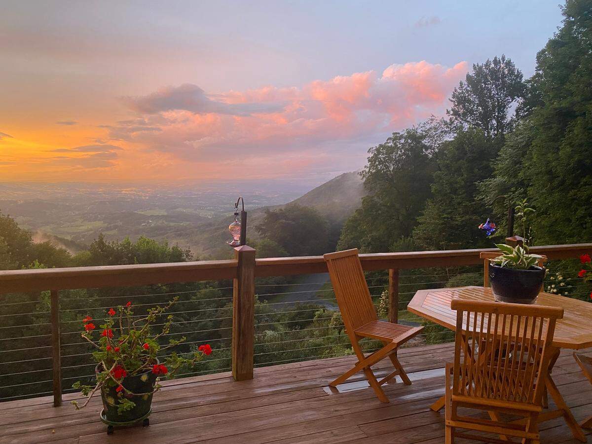 From the foreground of a wooden deck with a patio set, the view looks out onto a magnificent sunset. The sky is filled with glowing orange and pink clouds above a sprawling, misty valley and distant mountains. Potted plants and a lantern decorate the deck.