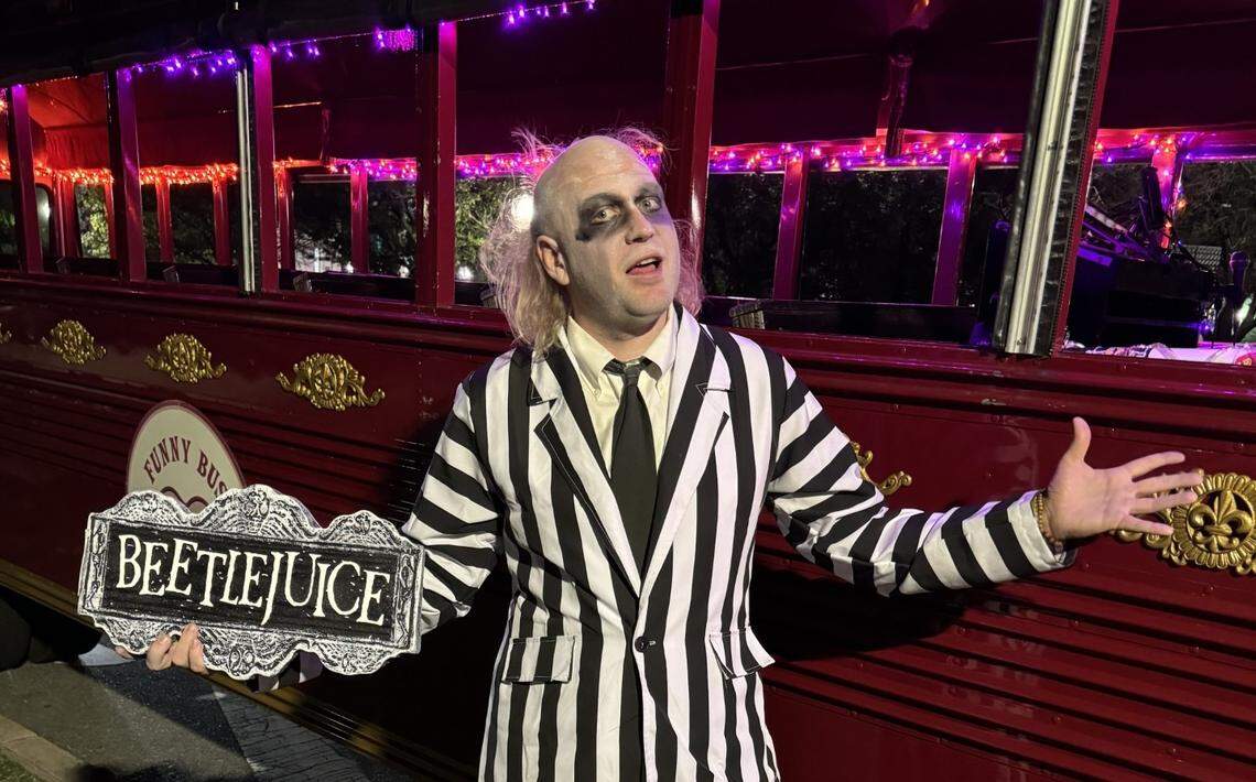 A performer in a full Beetlejuice costume, with a black-and-white striped suit and ghoulish makeup, poses dramatically inside a vehicle. The interior is bathed in purple light, and a glowing purple sign that reads “BEETLEJUICE” is visible in front of them.