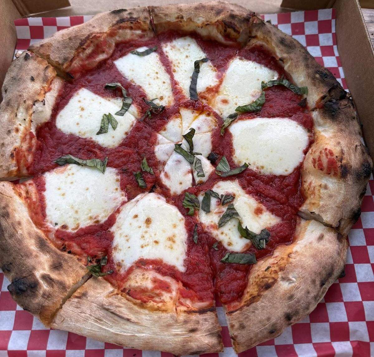 An overhead shot captures a sliced, artisanal Margherita pizza. The high-temperature bake is evident in the blistered and charred puffy crust. The pizza is topped with a simple red sauce, large ovals of melted fresh mozzarella, and garnished with fresh basil. The pizza rests on checkered paper inside a takeout box.