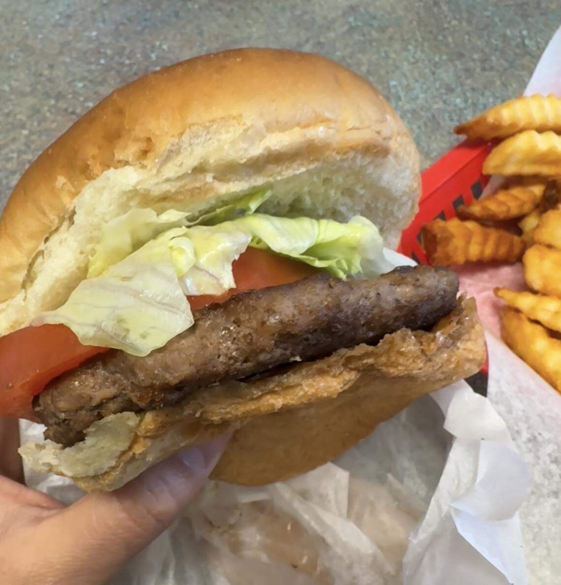 A handheld view of a burger with a thick patty, fresh lettuce and tomato on a large bun, accompanied by crinkle-cut fries in a red paper basket.