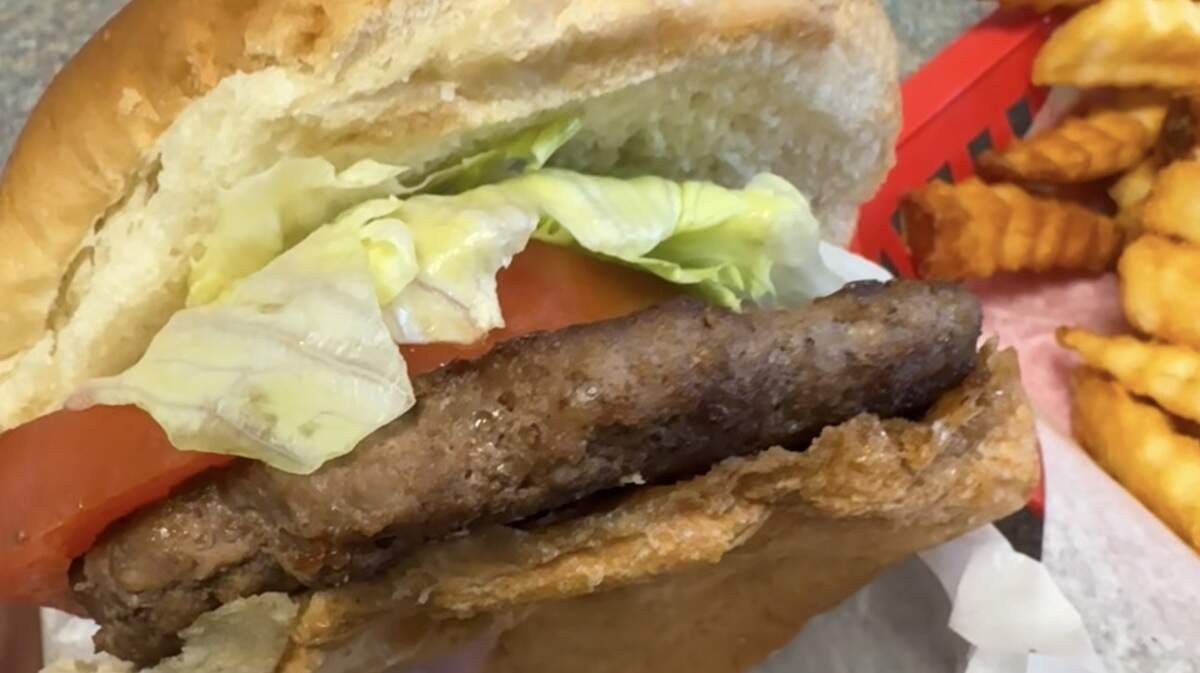 A handheld view of a burger with a thick patty, fresh lettuce and tomato on a large bun, accompanied by crinkle-cut fries in a red paper basket.
