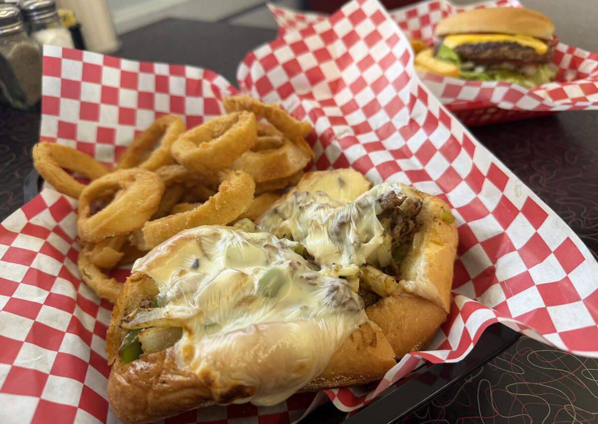 A close-up shot of a Philly cheesesteak sandwich on a toasted hoagie roll, covered in melted white cheese, sautéed peppers, and onions. It is served in a black plastic basket with a large side of crispy, breaded onion rings and lined with red and white checkered paper.