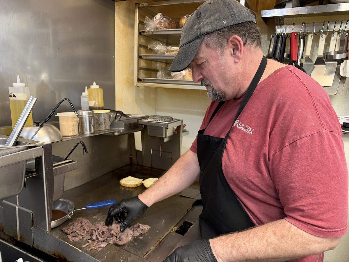 profile view of a chef wearing a grey baseball cap, red t-shirt, and black apron working in a professional kitchen, wearing black gloves and using one hand to spread out a large portion of thinly sliced roast beef on a hot flat-top grill. In the background, toasted buns sit on the griddle, and kitchen shelves hold various sauces, spices, and bread.