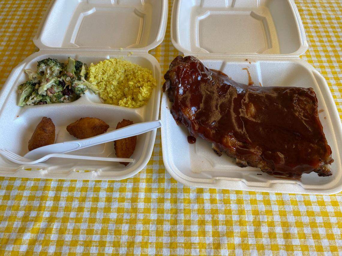 Two open white Styrofoam containers are side-by-side on a yellow and white checkered tablecloth. The Right Container contains a large rack of ribs generously covered in a glossy, dark barbecue sauce. The left container Contains several sides, including a broccoli salad with creamy dressing in the top-left, yellow coleslaw in the top-right, and three hushpuppies in the bottom section. A white plastic fork and knife are placed in the bottom section of the container with the sides.