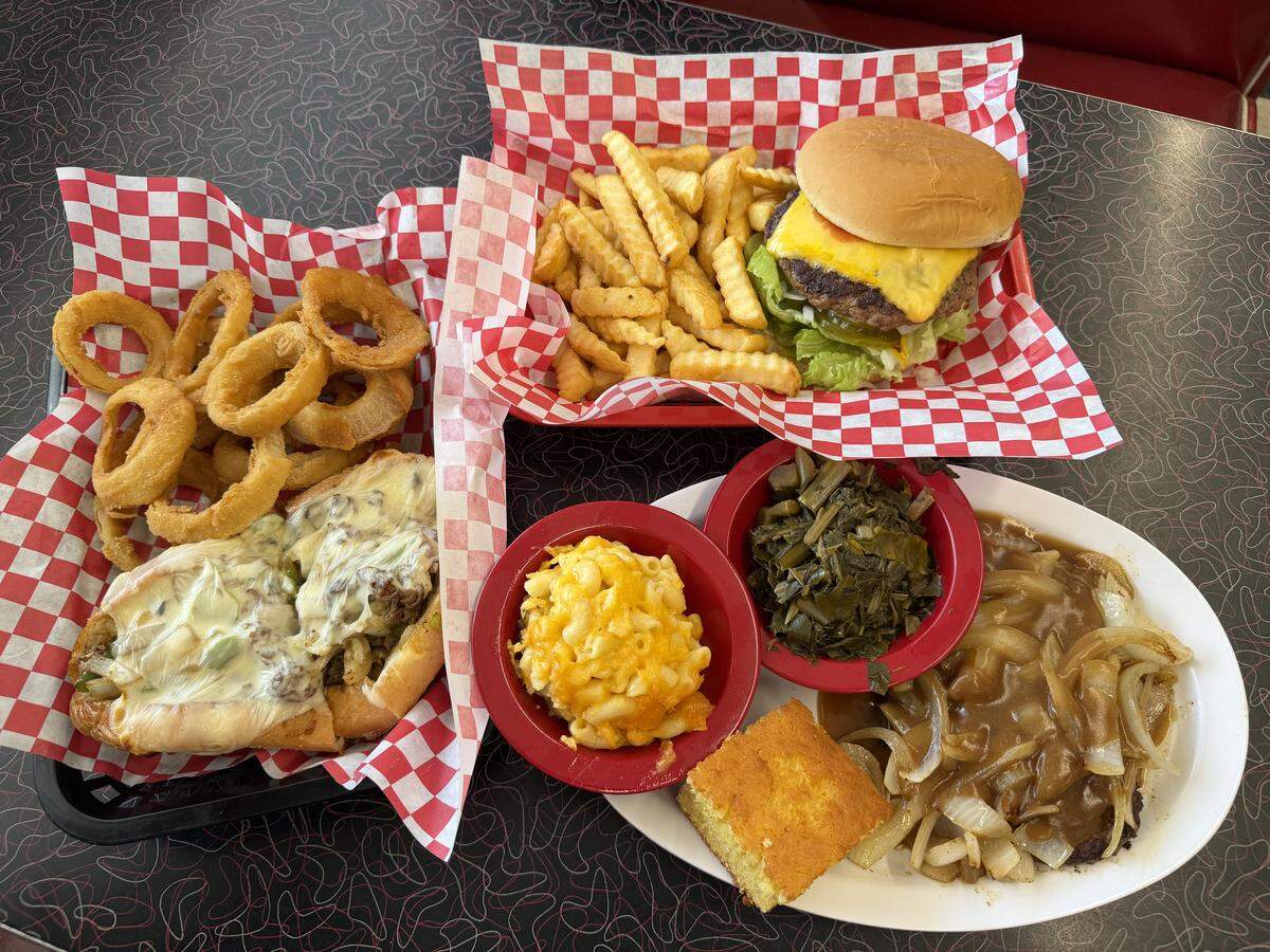 A top-down view of three full meal baskets on a retro patterned diner table. The spread includes a cheeseburger with crinkle-cut fries, a Philly cheesesteak sub with a large side of golden-brown onion rings, and a Southern platter featuring a hamburger steak smothered in gravy and onions, a square of cornbread, macaroni and cheese, and collard greens. All baskets are lined with red and white checkered paper.