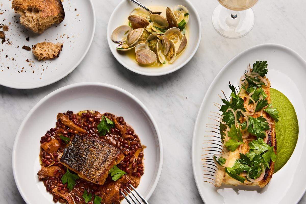 A bright, overhead flat lay shot of several gourmet dishes on a white marble table. In the bottom left, a large white bowl contains a seared fish fillet with crispy skin on a bed of dark grains and mushrooms. To the right, an oval plate holds another fish dish with a scored top, fresh herbs, and a vibrant green purée. At the top of the image are a small bowl of steamed clams in a light broth, a plate with remnants of crusty bread, and the top of a wine glass.