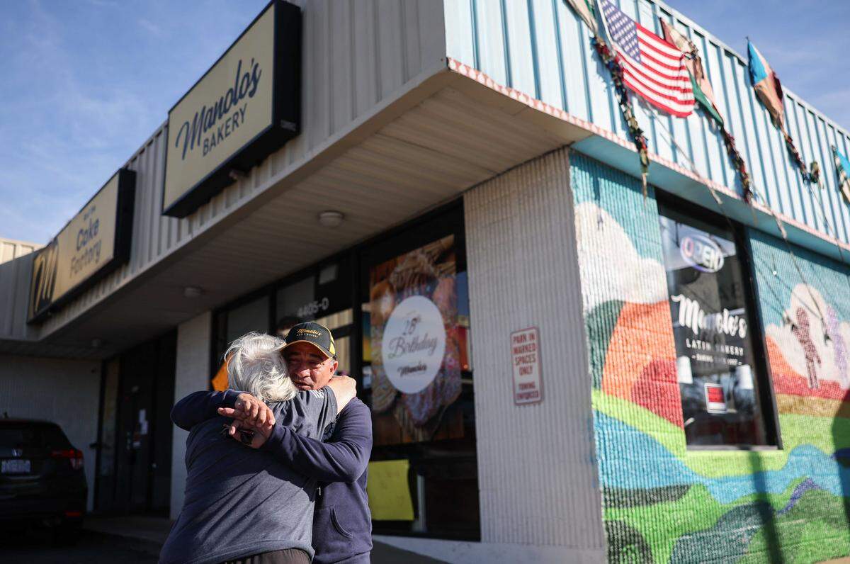 A slightly low-angle shot of a bakery owner and customer embracing tightly in front of Manolo’s Bakery. The bakery’s facade features a sign reading “Manolo’s BAKERY,” a large colorful mural on the windows, and an American flag hanging above.
