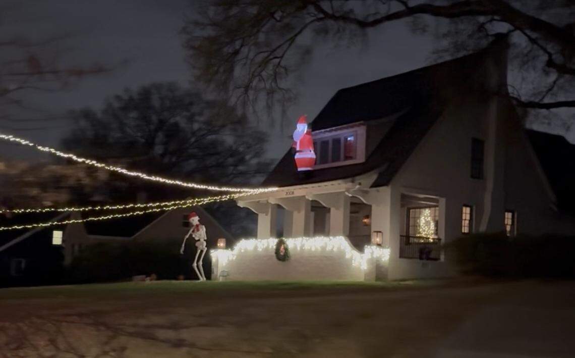 A dark, evening photo of a festive and humorous holiday display. The main house is decorated with white string lights, and an inflatable Santa is perched in the attic window. On the front porch, a life-sized skeleton figure, also wearing a Santa hat, stands near a small wreath, blending Halloween and Christmas themes.