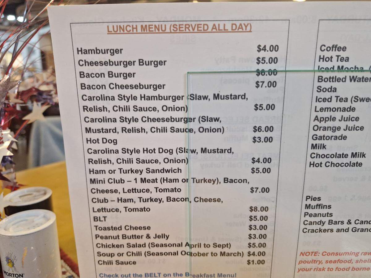 A close-up, high-angle shot of a printed “LUNCH MENU (SERVED ALL DAY)” displayed on a tabletop. The menu is divided into a left column with food items and a right column for beverages. The food section lists items like “Hamburger,” “Cheeseburger Burger,” “Bacon Burger,” and several “Carolina Style” options with slaw, mustard, relish, chili sauce, and onion. Prices are listed to the right of each item, with most ranging from $3.00 to $8.00. The right side of the menu shows drinks like “Coffee,” “Hot Tea,” and “Bottled Water,” and includes a warning about consuming raw or undercooked foods. The background is slightly blurred and shows some metallic containers.