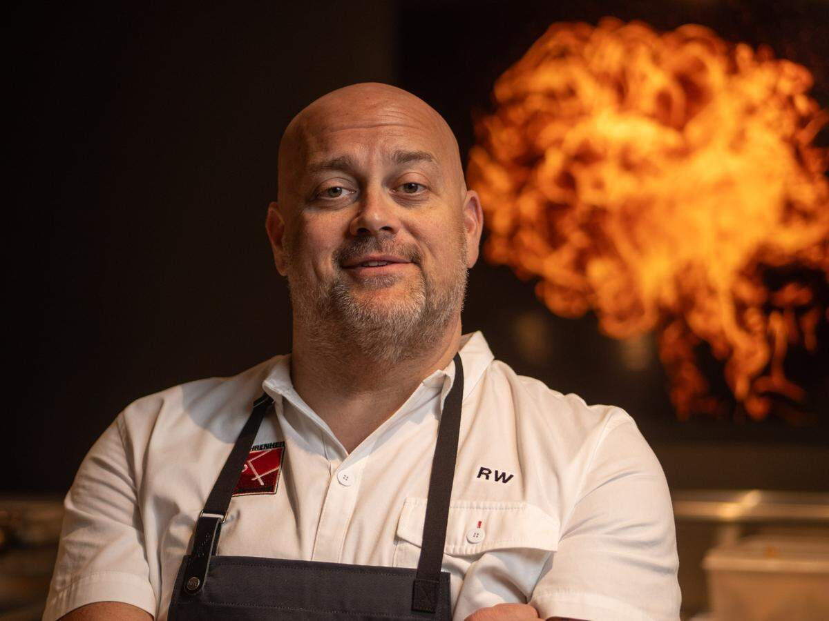 A bald man with a light beard and a faint smile is looking directly at the camera. He is wearing a white chef’s coat with an embroidered name or initials “RW” on the right chest and a dark apron. A red logo is visible on the left side of his chest. In the blurred background, a dramatic burst of orange flames erupts, likely from a kitchen grill or a similar cooking element, creating a striking contrast.