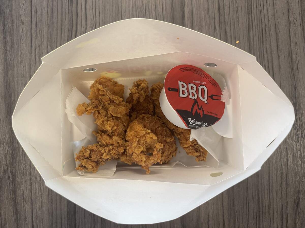A top-down view of an open white paperboard fast-food container sitting on a dark wooden table. Inside the box are several heavily breaded, crispy golden-brown chicken tenders resting on white greaseproof paper. A small, round Bojangles brand dipping sauce container with a red lid labeled “BBQ” is tucked into the corner next to the chicken.